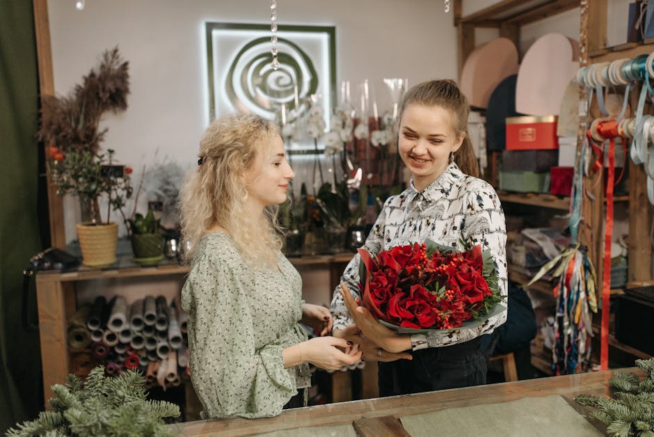 A florist at a flower shop called floristisleworth.co.uk presents a vibrant floral gift consisting of red roses and other red blooms, arranged with green foliage, wrapped in decorative paper and held 