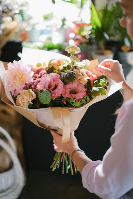A person holding a large bouquet of fresh flowers with pink and white blooms, including peonies, hydrangeas, and chrysanthemums, wrapped in cream-colored paper with a satin ribbon. The arrangement fea