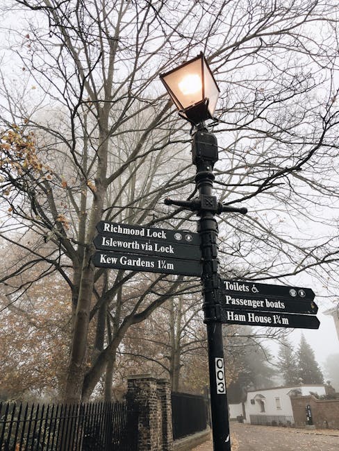 A close-up of a classic black street lamp post with a warm light glowing, situated outdoors among leafless trees on a foggy day. Attached to the post are three black directional signs indicating nearb