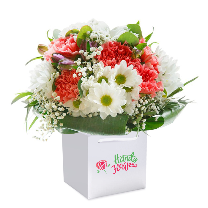 Resting in a refined square white gift bag, a vibrant, show-stopping bouquet is arranged with the grace and skill befitting long-standing Isleworth floristry. The assemblage of coral and deep-red carnations, their lavishly ruffled petals layered atop one another, radiates energy and affection-a nod to the spirited blooms found in private gardens along Church Street. Scattered through the arrangement, open-faced white daisies with lively yellow-green centres inject a burst of happiness, each petal crisp and full of life. Soft clouds of baby's breath light up the bouquet's heart, their fragile blossoms reminiscent of a gentle spring morning in Isleworth's parks. The visual texture is further enriched by the addition of a few closed, purple-kissed buds tucked along the left edge, their subtle hue promising growth and mystery. Rich, glossy green leaves cradle the stems, creating a secure, natural base that mirrors the winding green spaces along the Thames Path. The overall effect is of a full, rounded creation that feels both structured and exuberant-a fresh vision designed for celebration, connection, and local pride. The white carrier bag, equipped with strong ribbon handles, serves not only as an elegant vessel but also as a nod to the convenience offered by local flower delivery services. Adorned with a succinct artisan logo, the bag underscores its origins in Isleworth's trusted floral community. Lit with gentle, even light and set against a flawless white background, every detail of this arrangement is highlighted, inviting thoughts of thoughtful gifting for birthdays, anniversaries, or simply to share beauty across Isleworth.