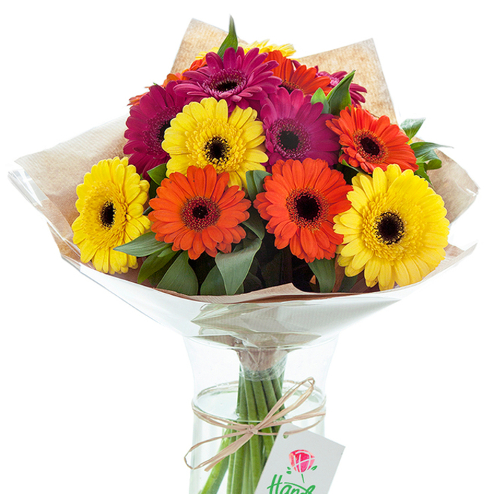 A richly textured bouquet of Gerbera daisies stands elegantly in a clear glass vase, the entire arrangement infused with the sunny optimism of Isleworth's lively market days. Each Gerbera-some golden yellow like morning sunlight on St John's Road, others burnt orange and magenta-radiates from its signature dark centre, creating an arresting play of contrast and harmony. The meticulous hand-tying, visible through the vase's surface, showcases the florist's detailed touch: every stem is neatly secured, their greens vibrant and crisp in the clear water below. Layers of natural-toned kraft paper provide a gently crinkled layer of warmth around the upper bouquet, blending seamlessly with shinier cellophane that both protects and celebrates the blossoms' sculptural form. The bouquet is completed with a simple twine bow, artfully knotted, from which hangs an understated gift tag printed with a stylised red rose and fragments of elegant lettering. This arrangement is more than just a burst of colour-it's a narrative of celebration, perfect for birthdays, anniversaries, or thoughtful surprises delivered anywhere in Isleworth, from historic Spring Grove to leafy Church Street. The sensory details-the cool, smooth glass, the soft rustle of paper, the bright hues against clean white-invite joy, gratitude, and that unmistakable feeling of giving or receiving something truly special from Florist Isleworth.